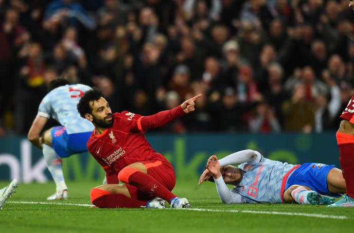 Mo Salah points behind him while half lying on his side with two other players lying next to him